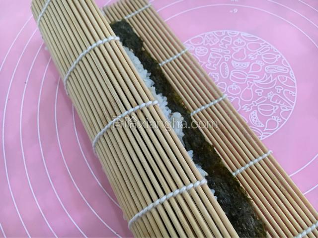 开始从下往上卷寿司帘，卷的时候注意不要把帘子卷进饭里。
边卷边向上拉拉帘子，每卷一圈都要用手握紧固定，卷到最后的地方，停下来在剩下的紫菜上面抹一点清水，再卷，这样紫菜就能贴的很紧也不会松散。
