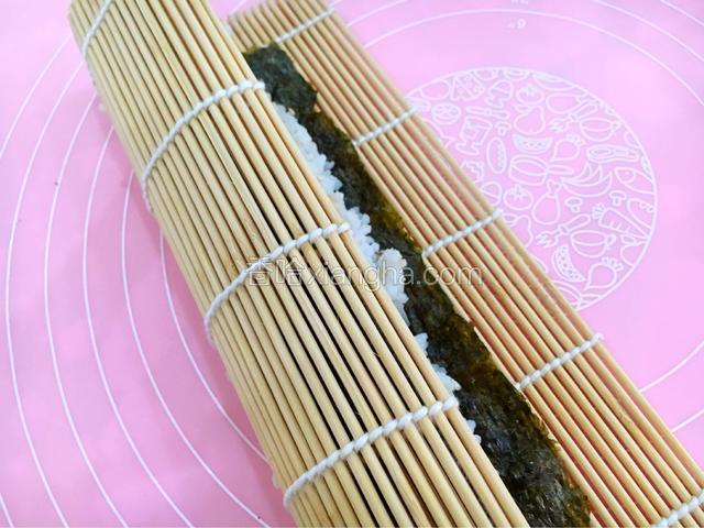 开始从下往上卷寿司帘，卷的时候注意不要把帘子卷进饭里。
边卷边向上拉拉帘子，每卷一圈都要用手握紧固定，卷到最后的地方，停下来在剩下的紫菜上面抹一点清水，再卷，这样紫菜就能贴的很紧也不会松散。