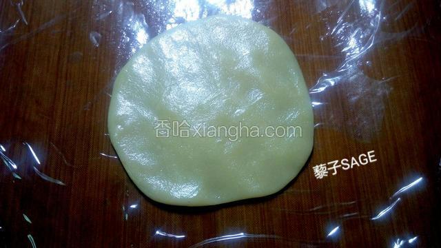取一个饼皮面团在保鲜膜上按扁，包入一颗白芸豆蛋黄馅。（饼皮面团比较粘手，在饼皮下面垫入一小张保鲜膜包裹起来会更方便。） 